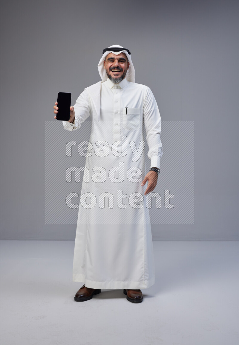 Saudi man Wearing Thob and white Shomag standing showing phone to camera on Gray background