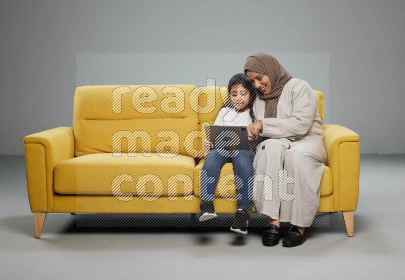 A girl with her mother on yellow sofa and watching on iPad on gray background