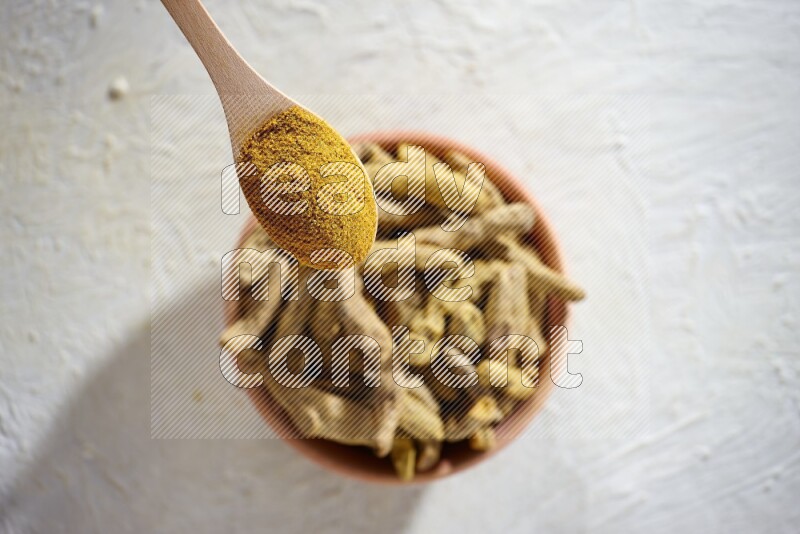 A wooden spoon full of turmeric powder above a wooden bowl full of dried turmeric whole fingers on a textured white flooring