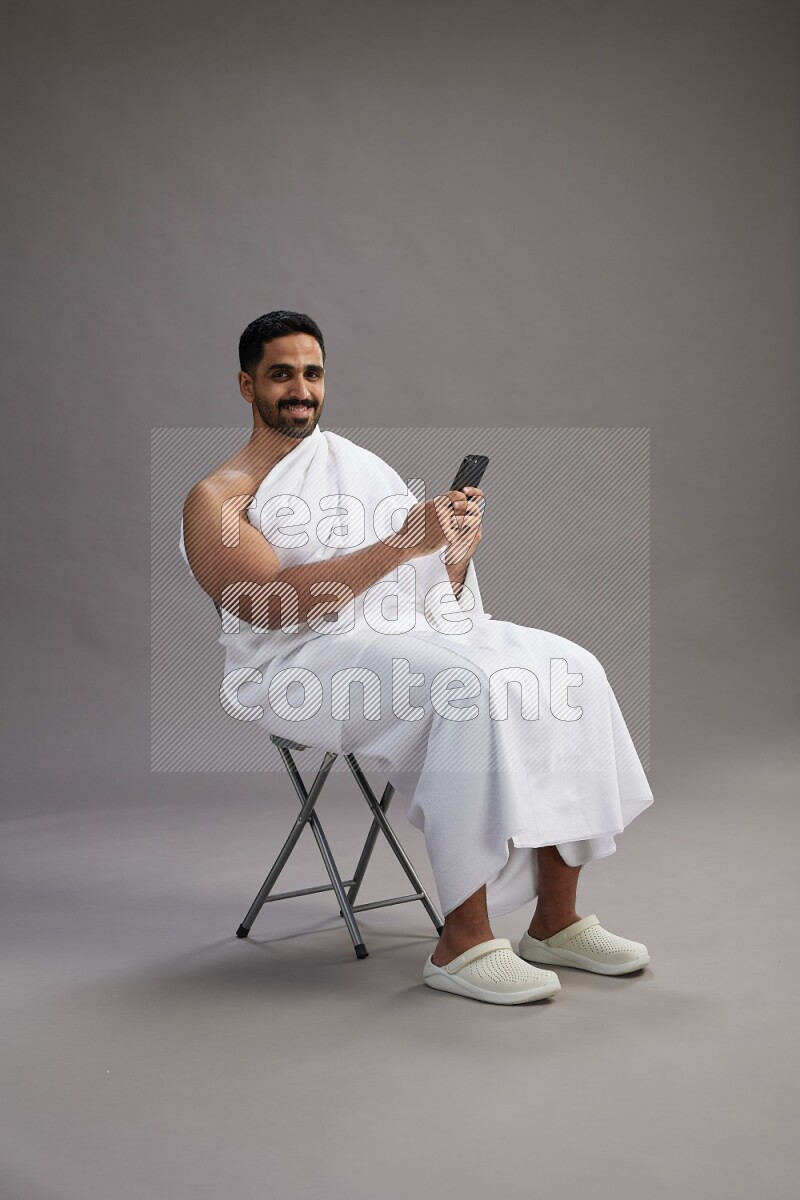 A man wearing Ehram sitting on chair texting on phone on gray background