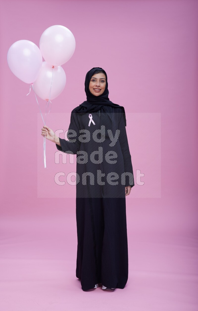 Saudi woman wearing pink ribbon on Abaya standing holding balloons on plain pink background