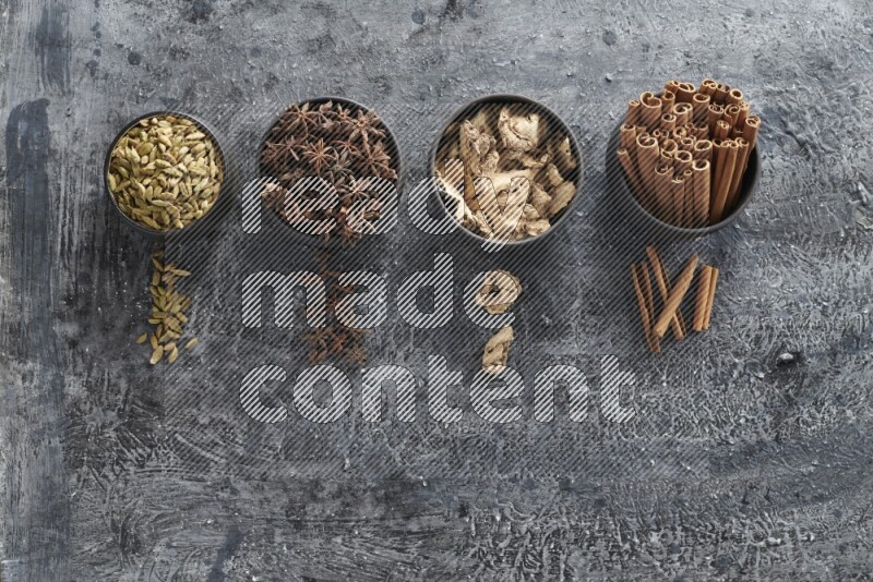 Ginger, Cardamom, Star anise and cinnamon sticks in 4 bowls on a textured black background