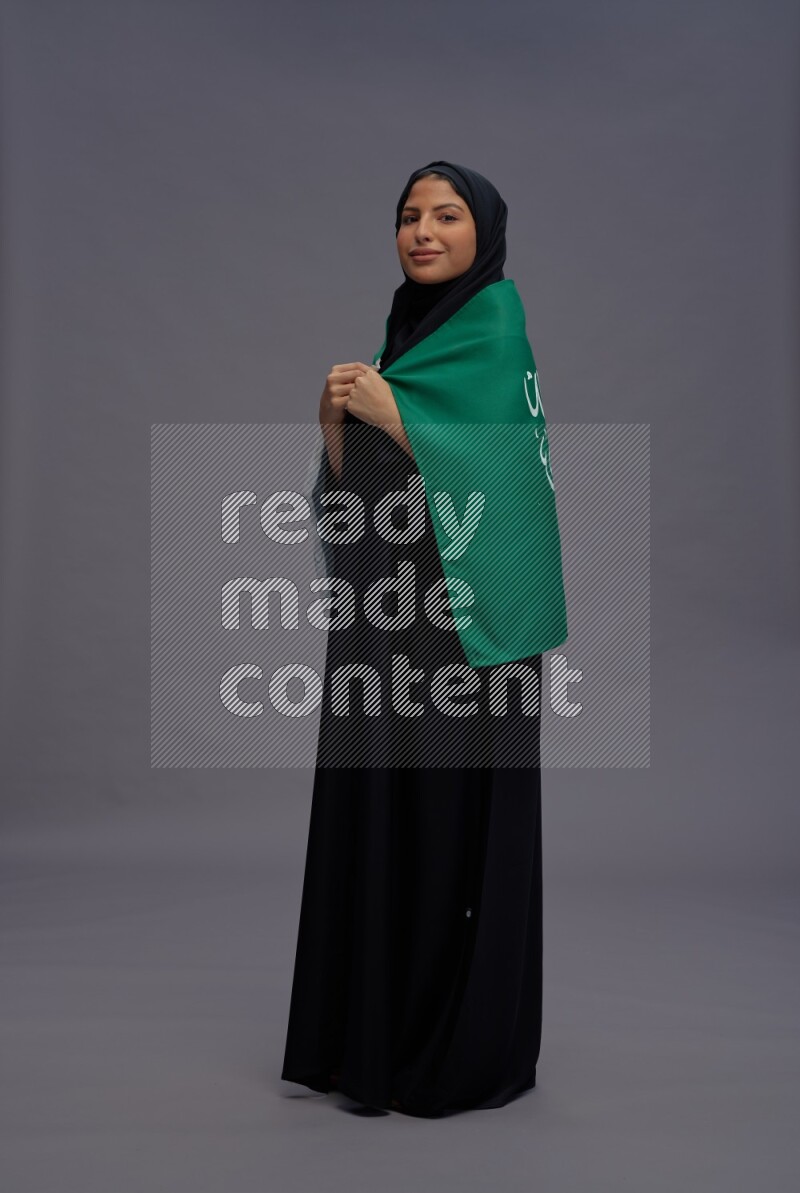 Saudi woman wearing Abaya standing holding Saudi flag on gray background