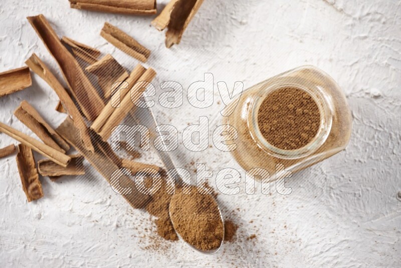 Herbal glass jar full cinnamon powder and a metal spoon surrounded by cinnamon sticks on a white background