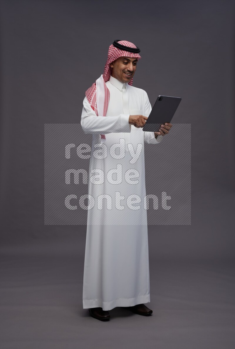 Saudi man wearing thob and shomag standing working on tablet on gray background