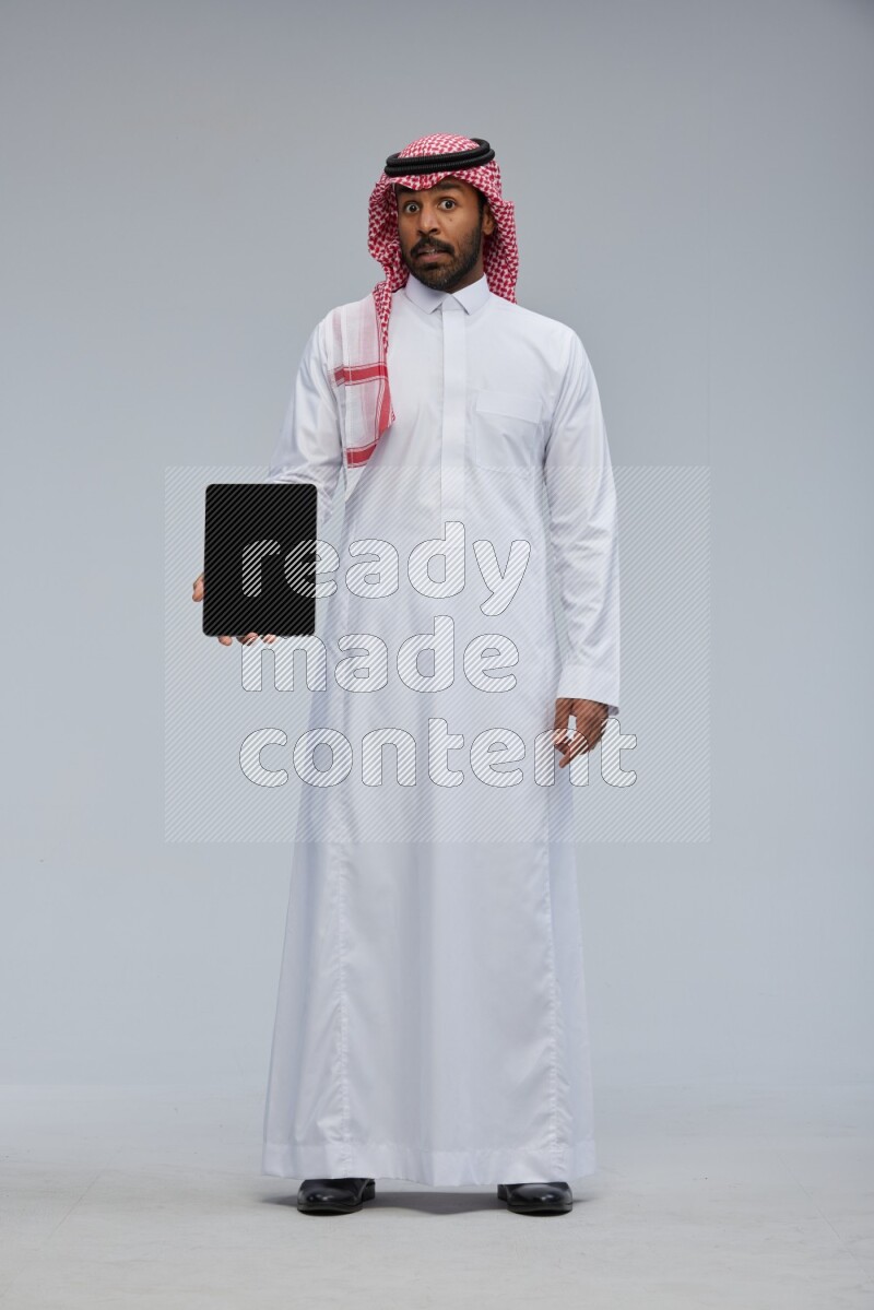 Saudi man Wearing Thob and shomag standing showing tablet to camera on Gray background