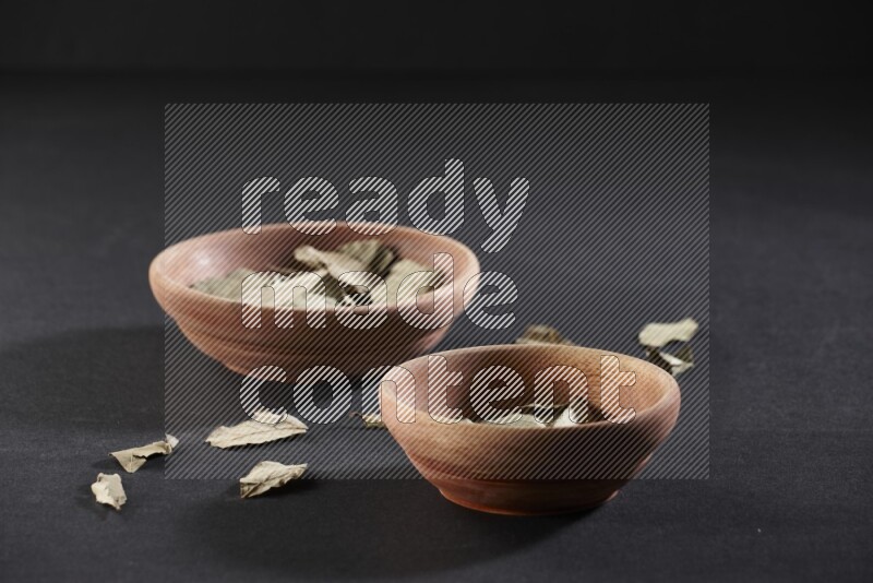2 wooden bowls full of dried bay leaves with more leaves spread on black flooring