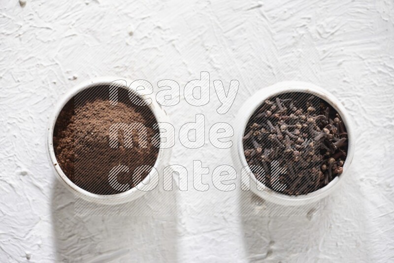 2 Beige ceramic bowls, one full of cloves and the other full of powder on textured white flooring