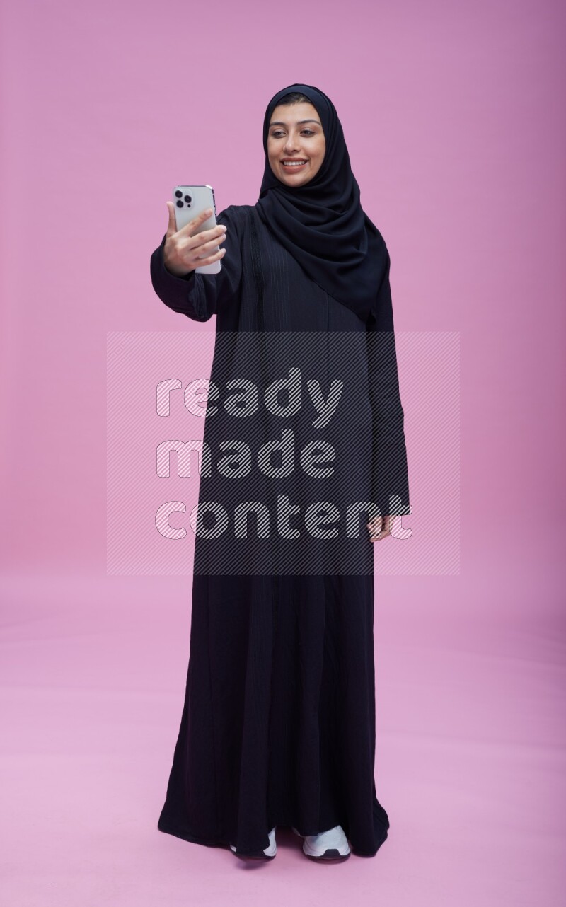 Saudi woman wearing Abaya standing taking selfie on pink background