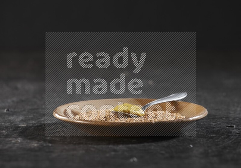 A beige pottery plate filled with mustard seeds with a metal spoon full of mustard paste in it on a textured black flooring