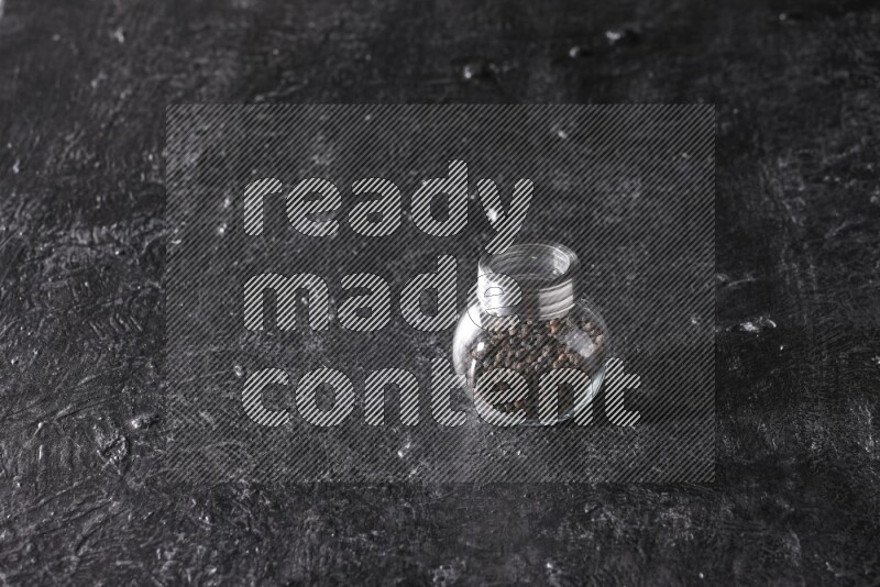 A glass spice jar full of black pepper beads on textured black flooring