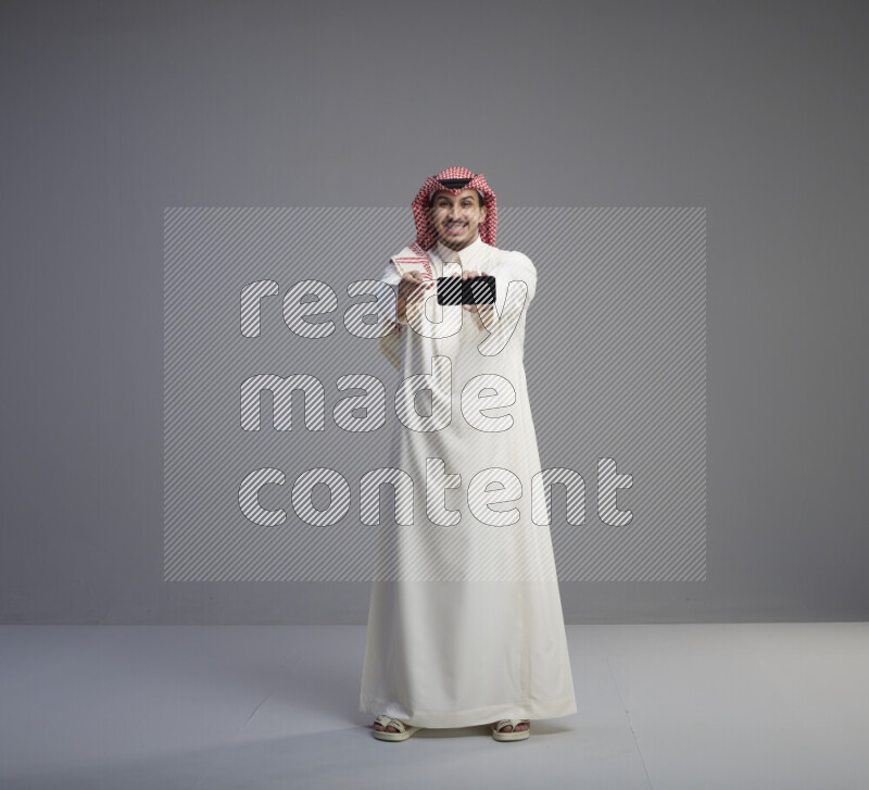 A Saudi man standing wearing thob and red shomag showing phone to camera on gray background
