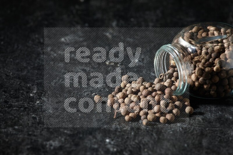 A flipped glass spice jar full of allspice whole balls and the balls spilled out of it on a textured black flooring