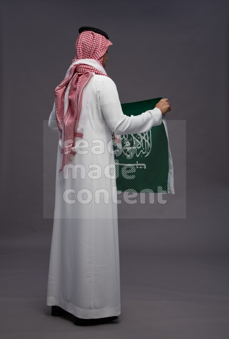 Saudi man wearing thob and shomag standing holding Saudi flag on gray background