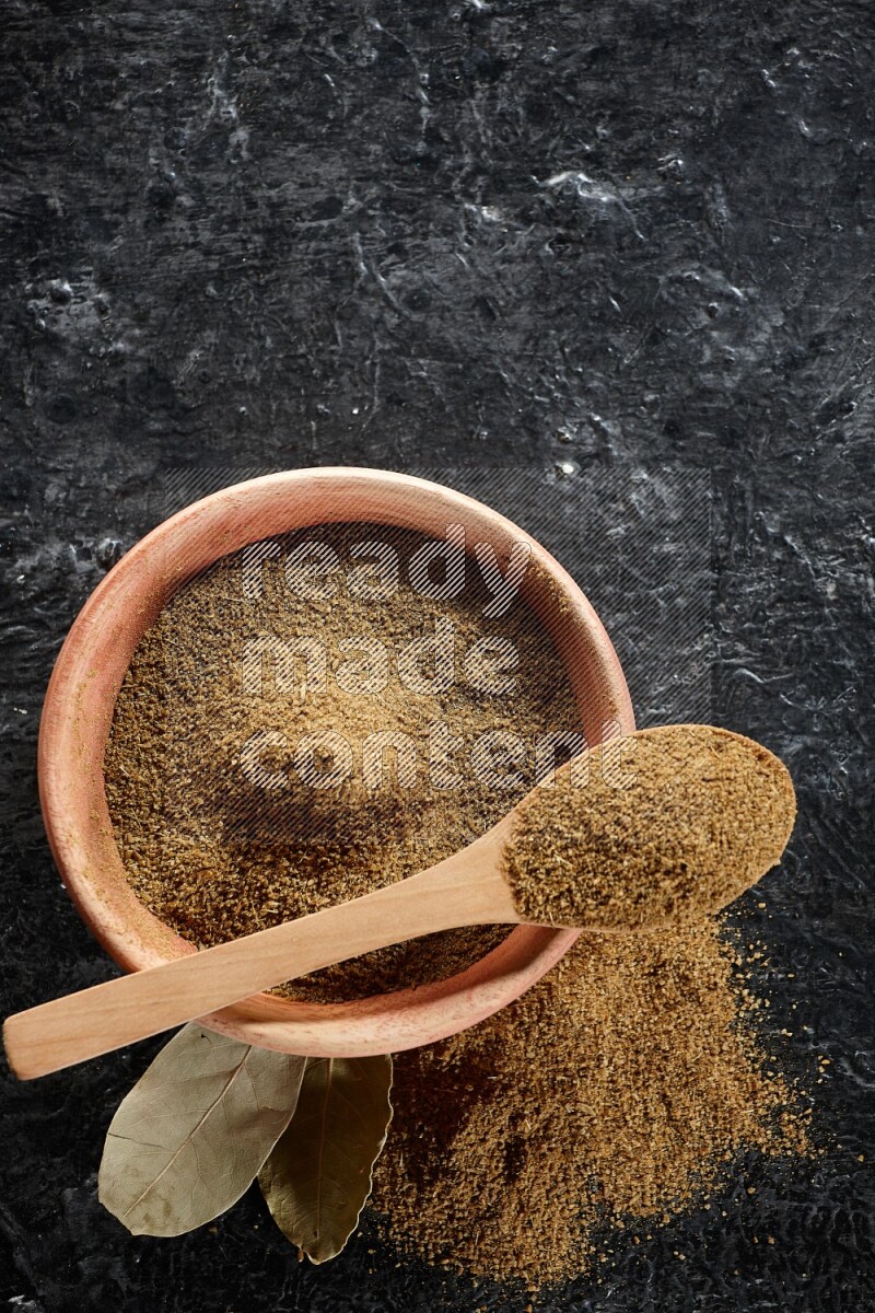 A wooden bowl and spoon full of cumin powder on a textured black flooring