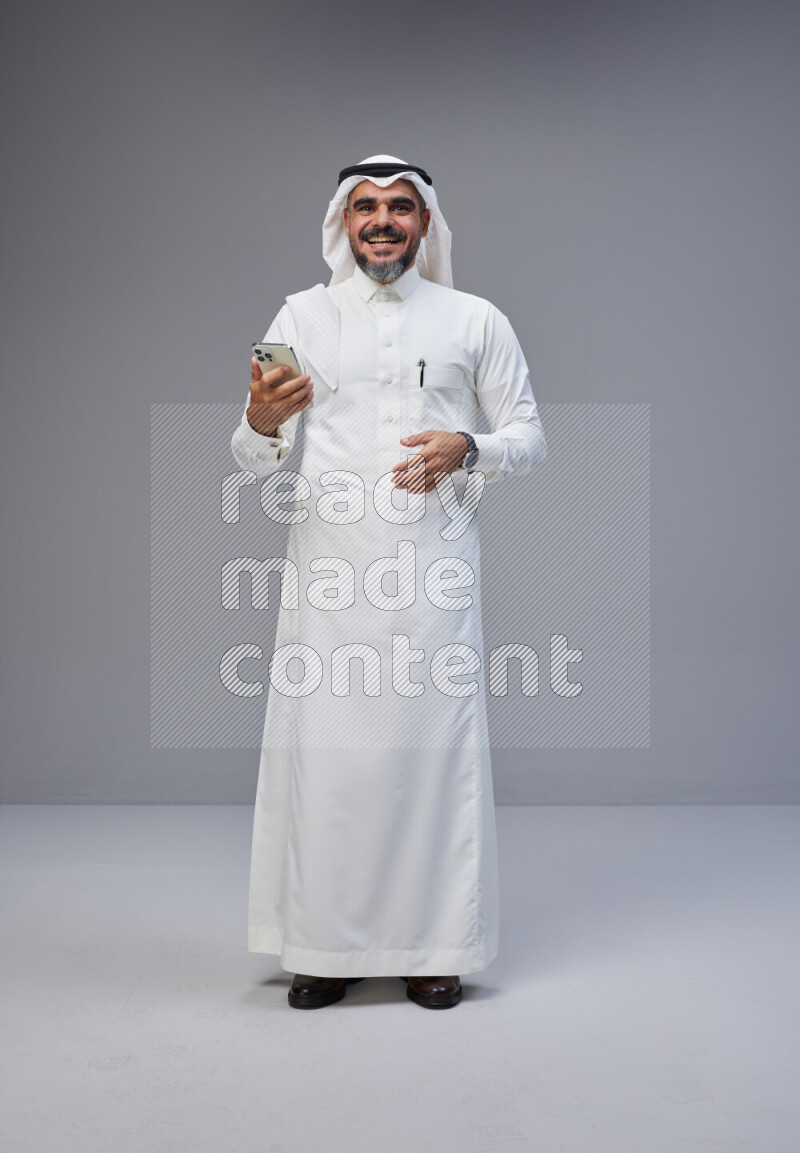 Saudi man Wearing Thob and white Shomag standing texting on phone on Gray background