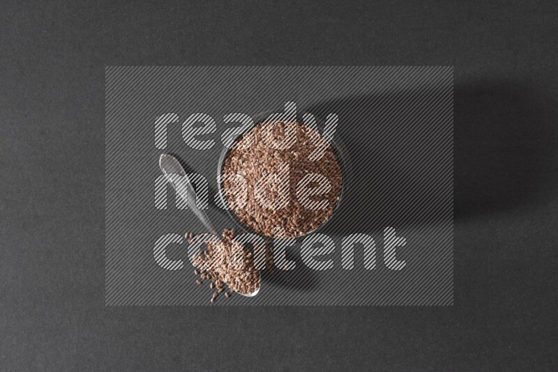 وعاء فخاري أسود ممتلئ بحبوب بذر الكتان مع ملعقة معدنية ممتلئة على أرضية سوداء