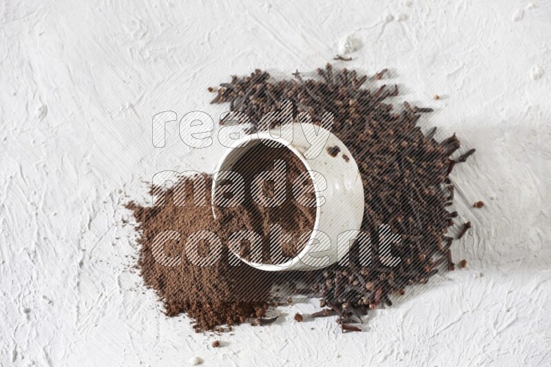 A beige ceramic bowl with cloves powder spilled out of it on a textured white flooring