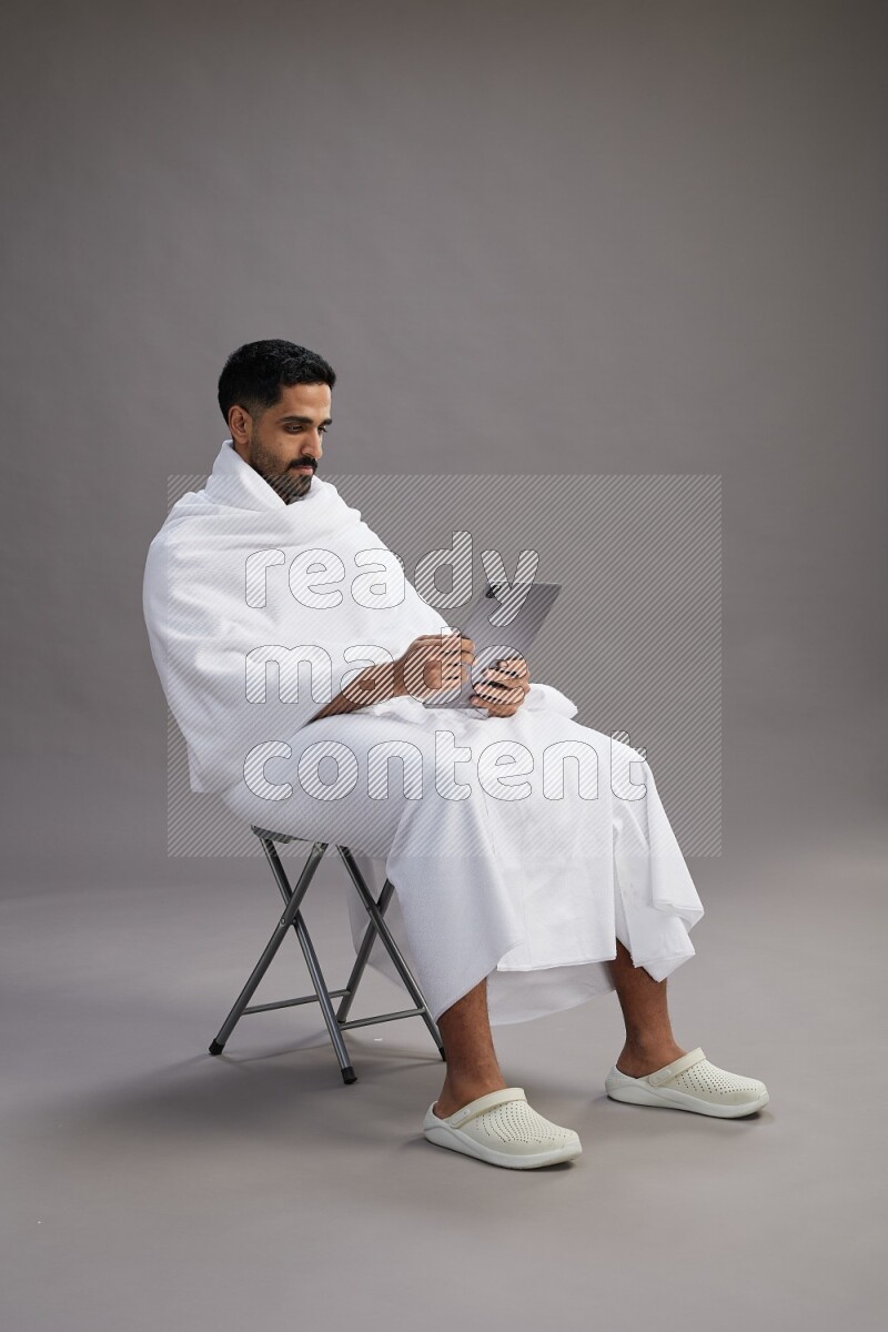 A man wearing Ehram sitting on chair holding a tablet on gray background