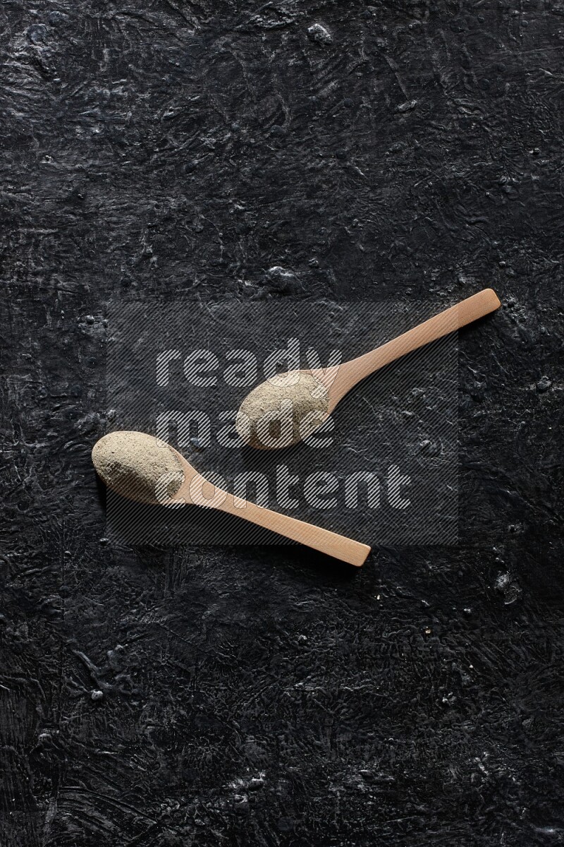2 wooden spoons full of white pepper powder on textured black flooring