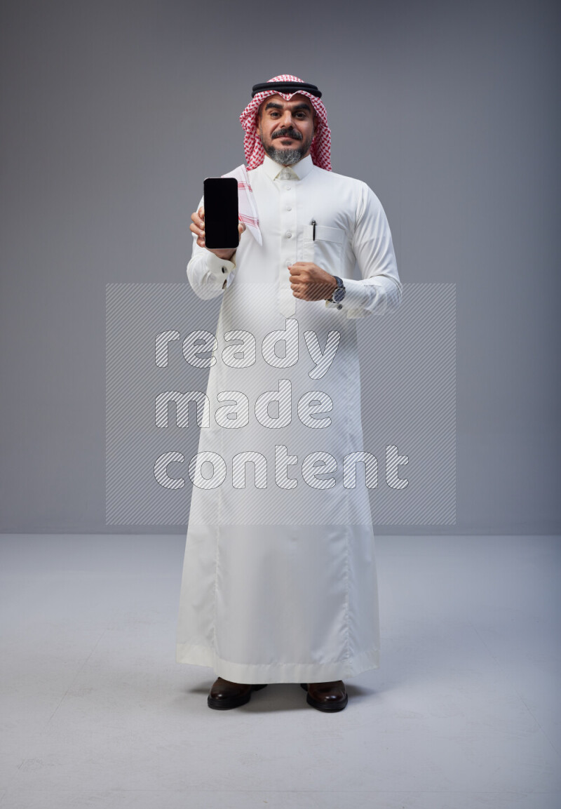 Saudi man Wearing Thob and red Shomag standing showing phone to camera on Gray background