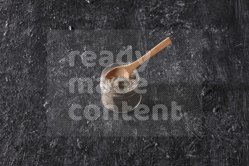 A glass jar full of black pepper powder and a wooden spoon on a textured black flooring