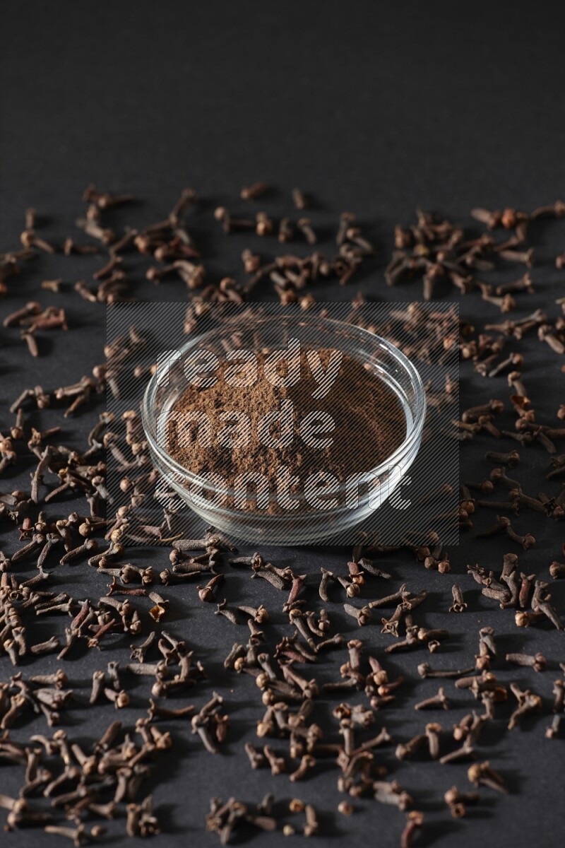 A glass bowl full of cloves powder with cloves grains spread on black flooring