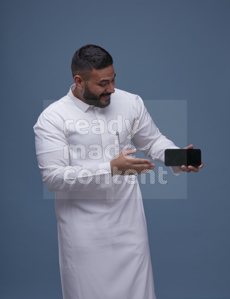 A man Showing his phone in Blue Background wearing Saudi Thob
