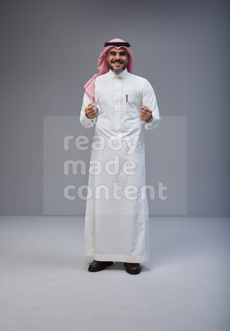 Saudi man Wearing Thob and red white Shomag standing interacting with the camera on Gray background