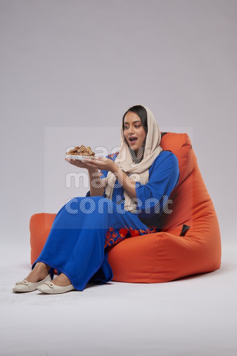 A Woman sitting on an orange beanbag wearing Jalabeya holding a plate of dates