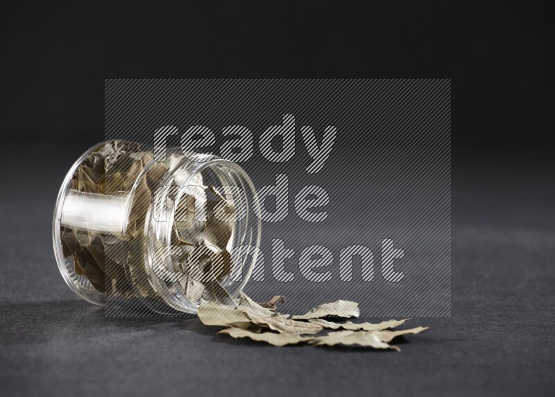 A glass jar full of dried bay leaves and jar is flipped and leaves spread out on black flooring