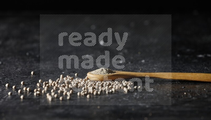 A wooden spoon full of white pepper powder with white pepper beads on textured black flooring