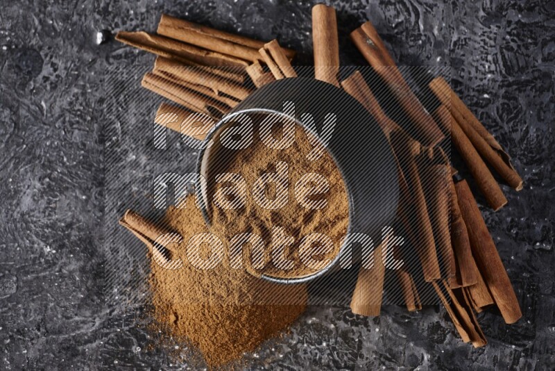 Black pottery bowl over filled with cinnamon powder and cinnamon sticks around the bowl on a textured black background
