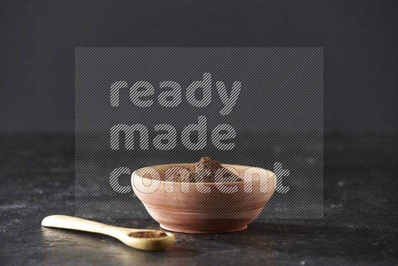 A wooden bowl and wooden spoon full of cloves powder on textured black flooring