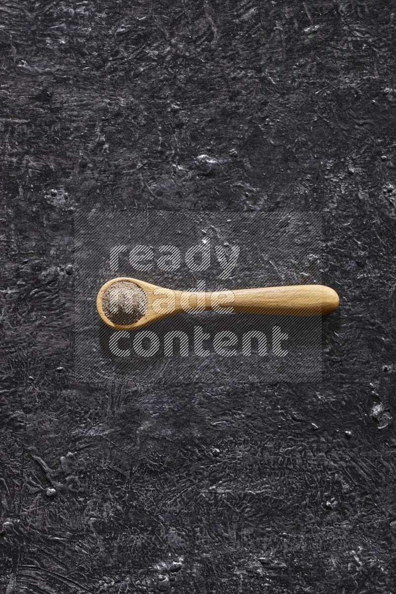 A wooden spoon full of black pepper powder on a textured black flooring