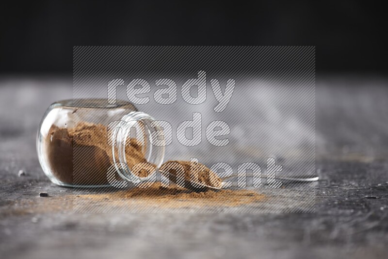 Herbal glass jar full of cinnamon powder flipped and a metal spoon on textured black background