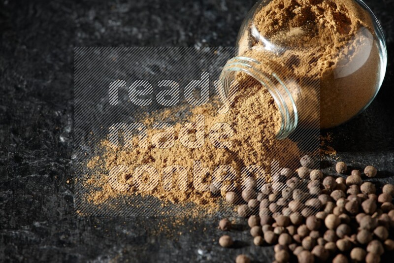 A flipped glass spice jar full of allspice powder and powder spilled out of it with whole balls on a textured black flooring