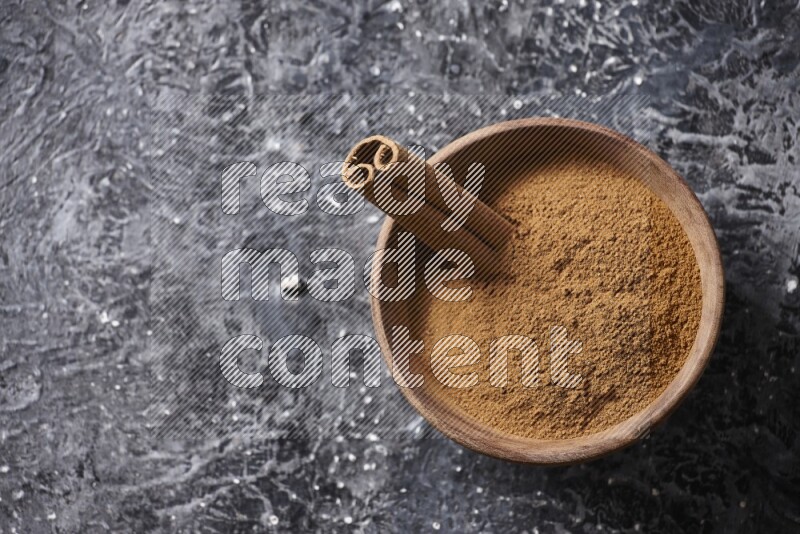 Wooden bowl full of cinnamon powder and a cinnamon stick on a textured black background