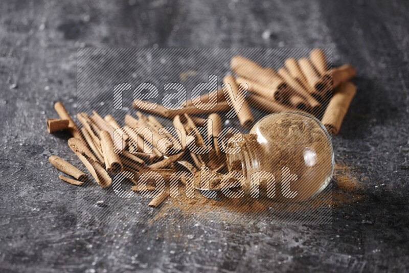 Herbal glass jar full cinnamon powder flipped and a metal spoon full of powder surrounded by cinnamon sticks on textured black background in different angles