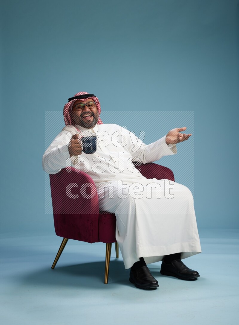 Saudi Man with shimag sitting on chair drinking coffee on blue background