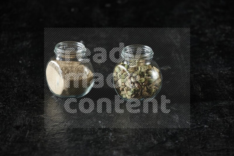 2 glass spice jars full of cardamom powder and cardamom seeds on textured black flooring
