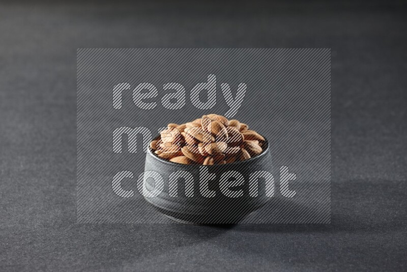 A black pottery bowl full of peeled almonds on a black background in different angles