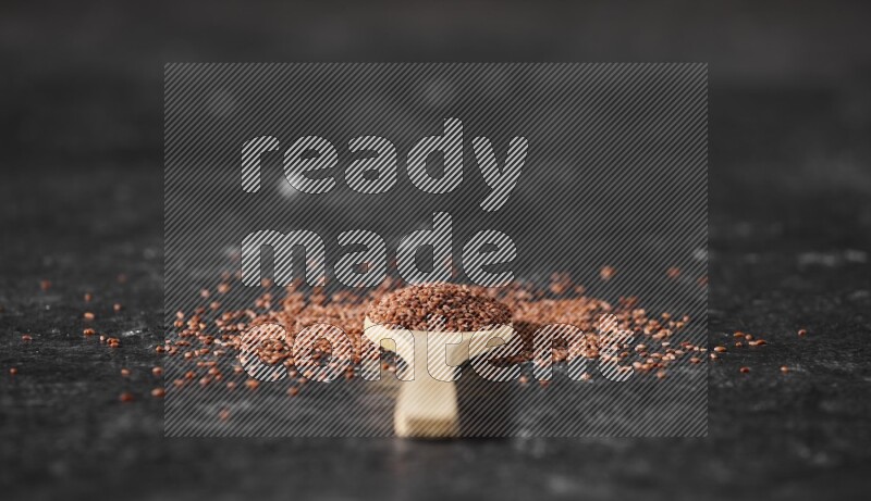 A wooden spoon full of garden cress seeds surrounded by the seeds on a textured black flooring