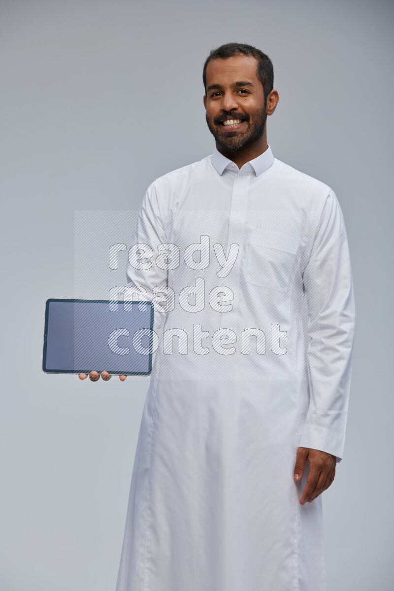 Saudi man wearing Thob standing showing tablet to camera on Gray background
