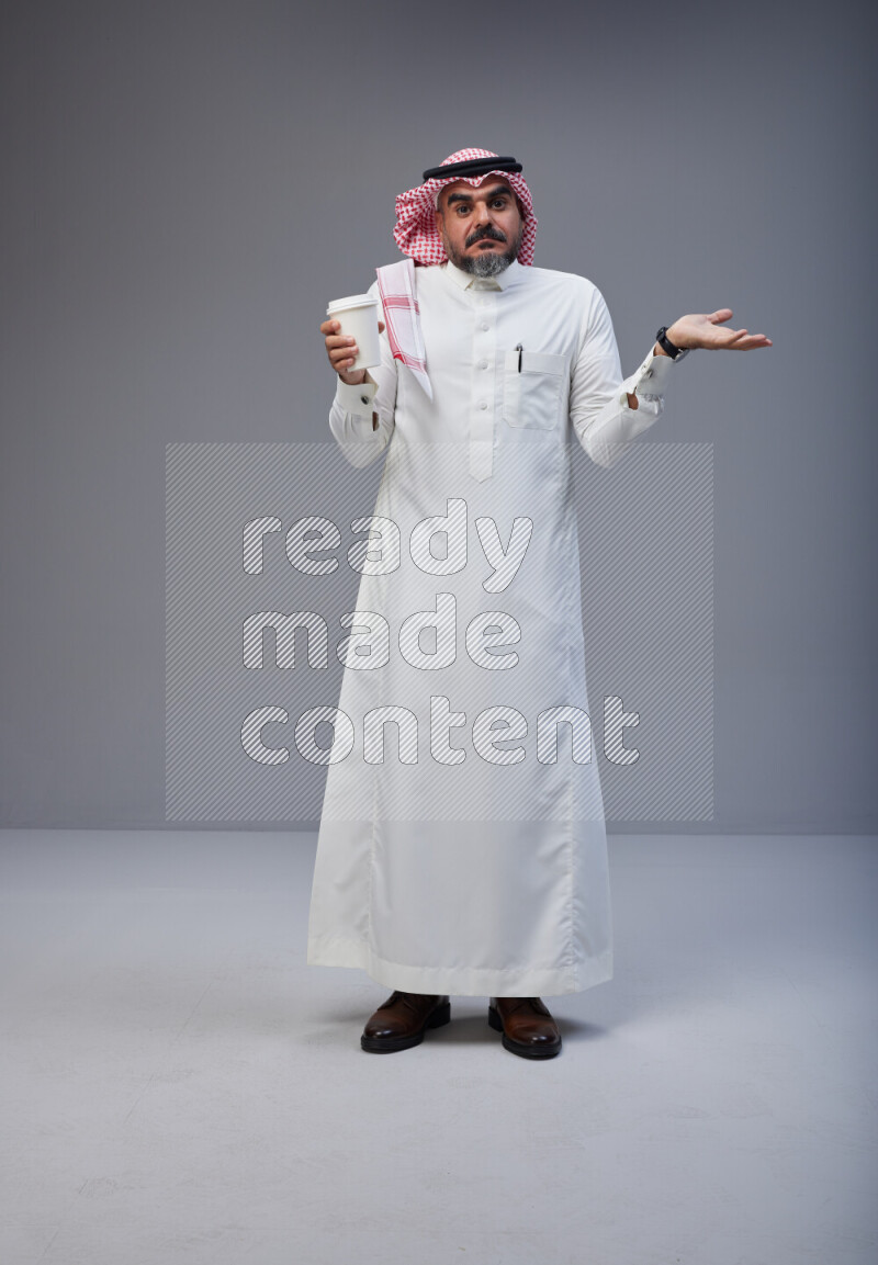 Saudi man Wearing Thob and red Shomag standing holding paper cup on Gray background