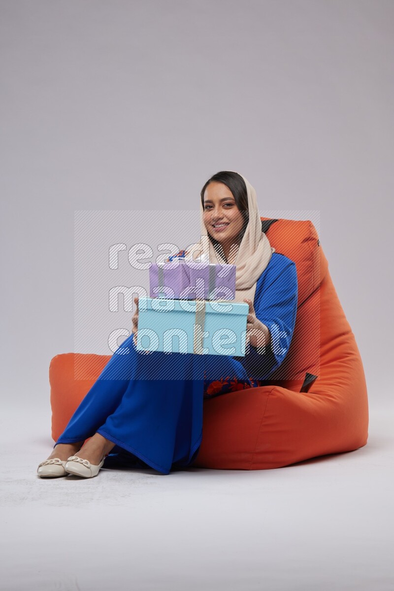 A woman sitting on an orange beanbag wearing Jalabeya holding a gift box