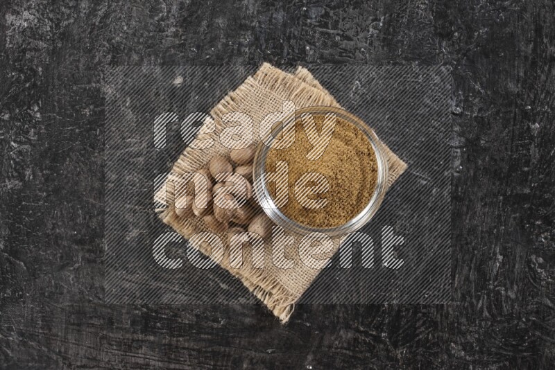 A glass bowl full of nutmeg powder with whole seeds beside it on burlap fabric on a textured black flooring