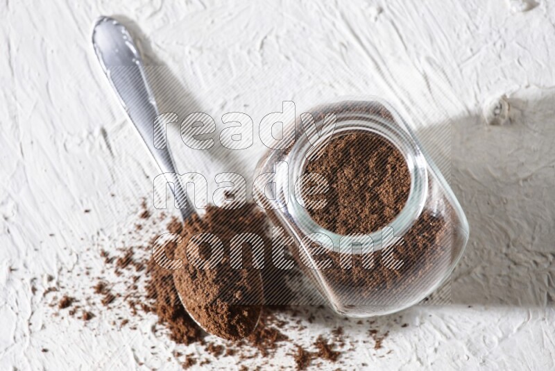 A glass spice jar and a metal spoon full of cloves powder on textured white flooring