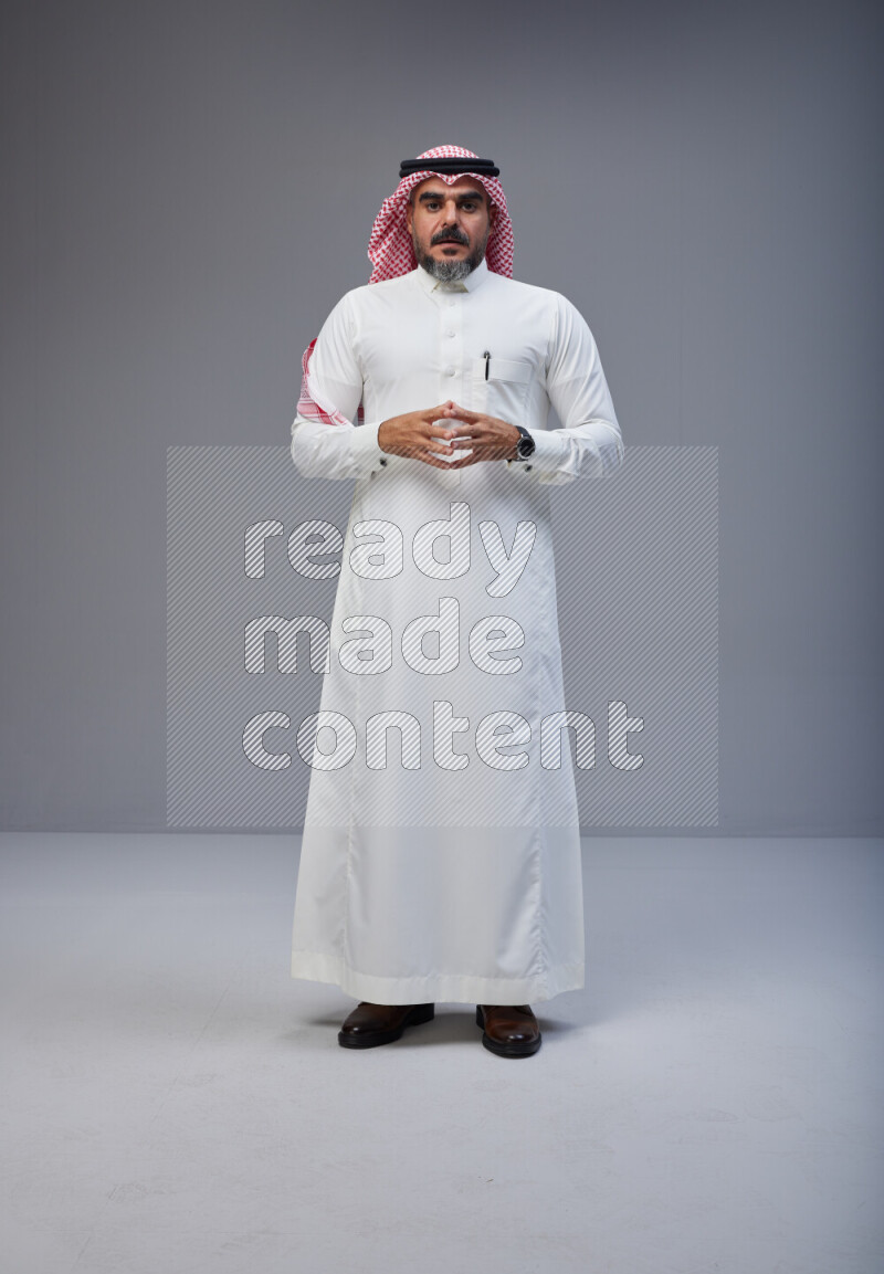 Saudi man Wearing Thob and red white Shomag standing interacting with the camera on Gray background