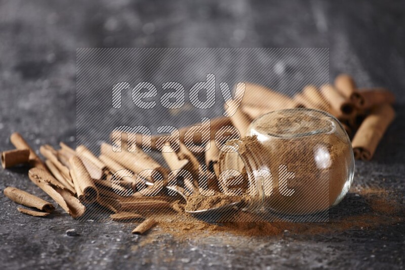 Herbal glass jar full cinnamon powder flipped and a metal spoon full of powder surrounded by cinnamon sticks on textured black background in different angles